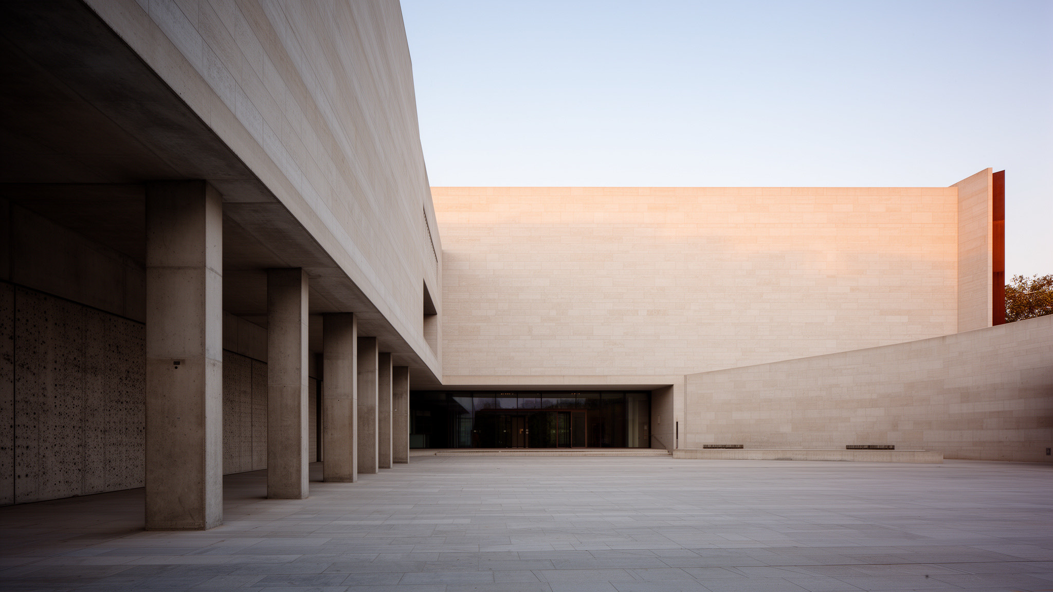 A quiet contemporary civic building in pale stone at early morning, long side light revealing its massing and a calm empty forecourt
